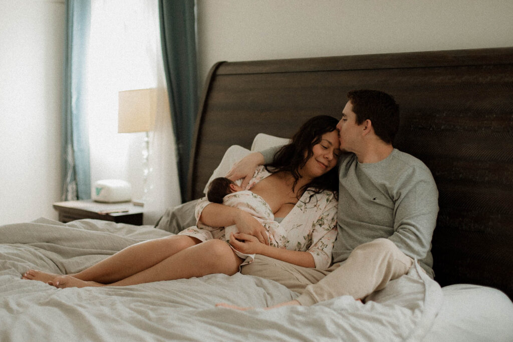 Mother nursing baby on bed with partner close beside her during cozy in-home newborn session