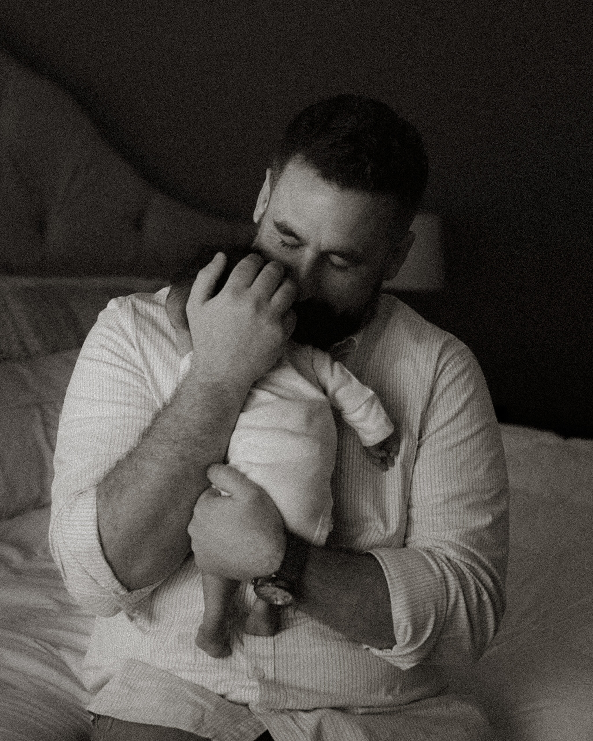 A father sits on a bed, eyes closed, holding his newborn daughter tightly against his chest. The image is in black and white, capturing a quiet and emotional moment of connection between father and baby.