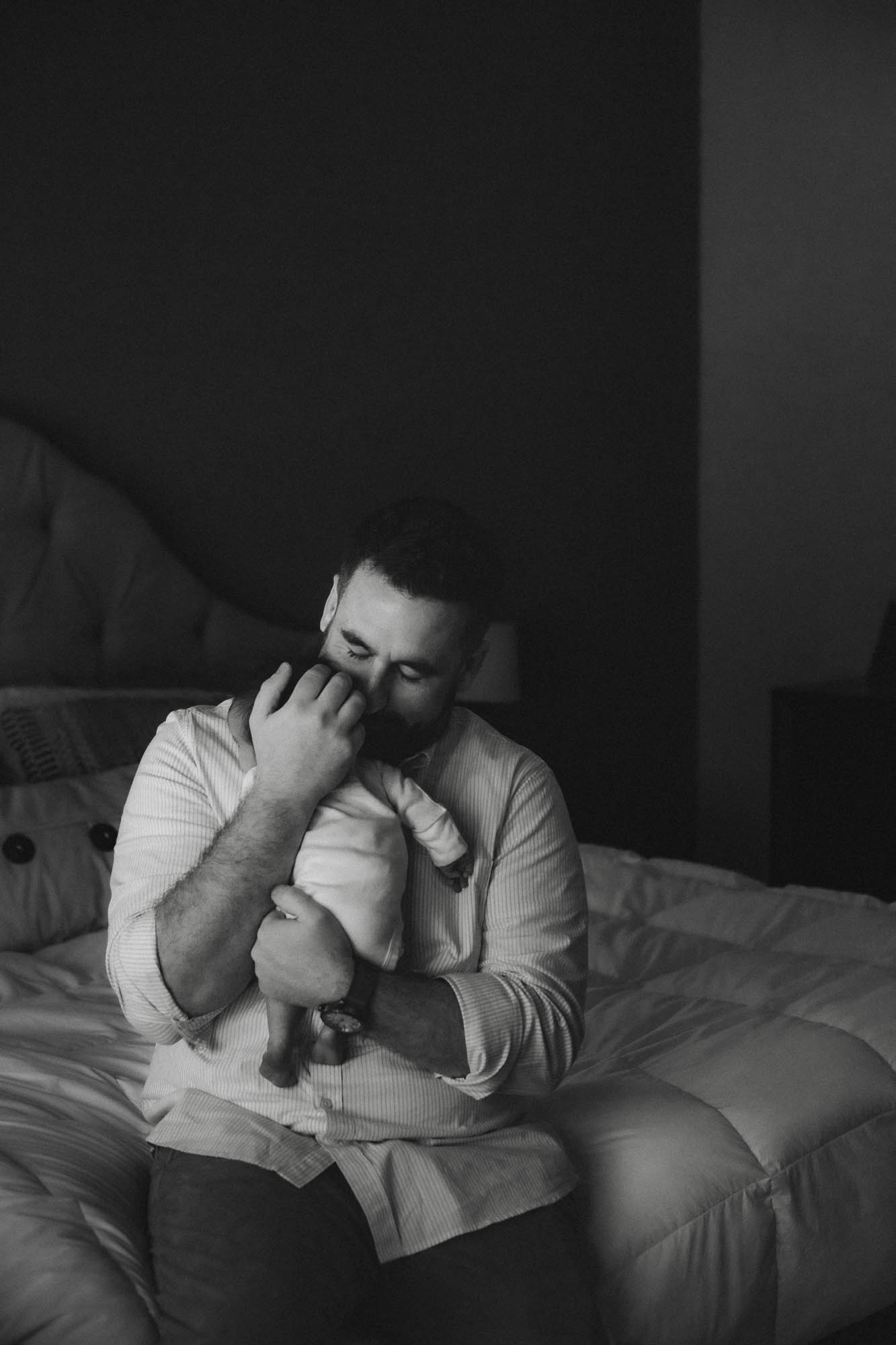 A father sits on a bed, eyes closed, holding his newborn daughter tightly against his chest. The image is in black and white, capturing a quiet and emotional moment of connection between father and baby